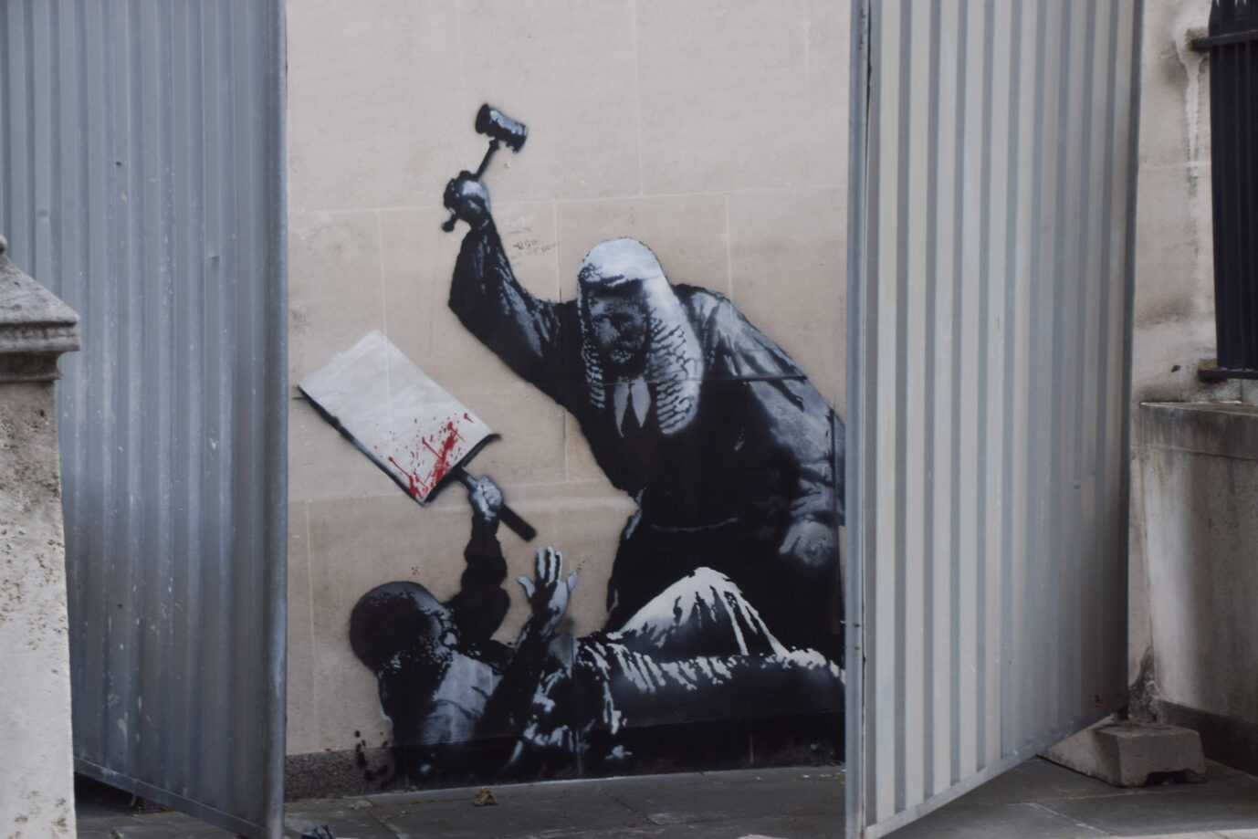 Banksys neues Gemälde: Mit Metallwänden verdeckt. Das Werk stellt einen Richter im traditionellen Gewand mit einer Allongeperücke dar, der einen auf dem Boden liegenden Protestierer zu erschlagen droht. Der Bedrohte hält ein blutüberströmtes Schild ohne Aufschrift. In Großbritannien findet derzeit eine Debatte über Zensur und Meinungsfreiheit statt. (Themenbild)
