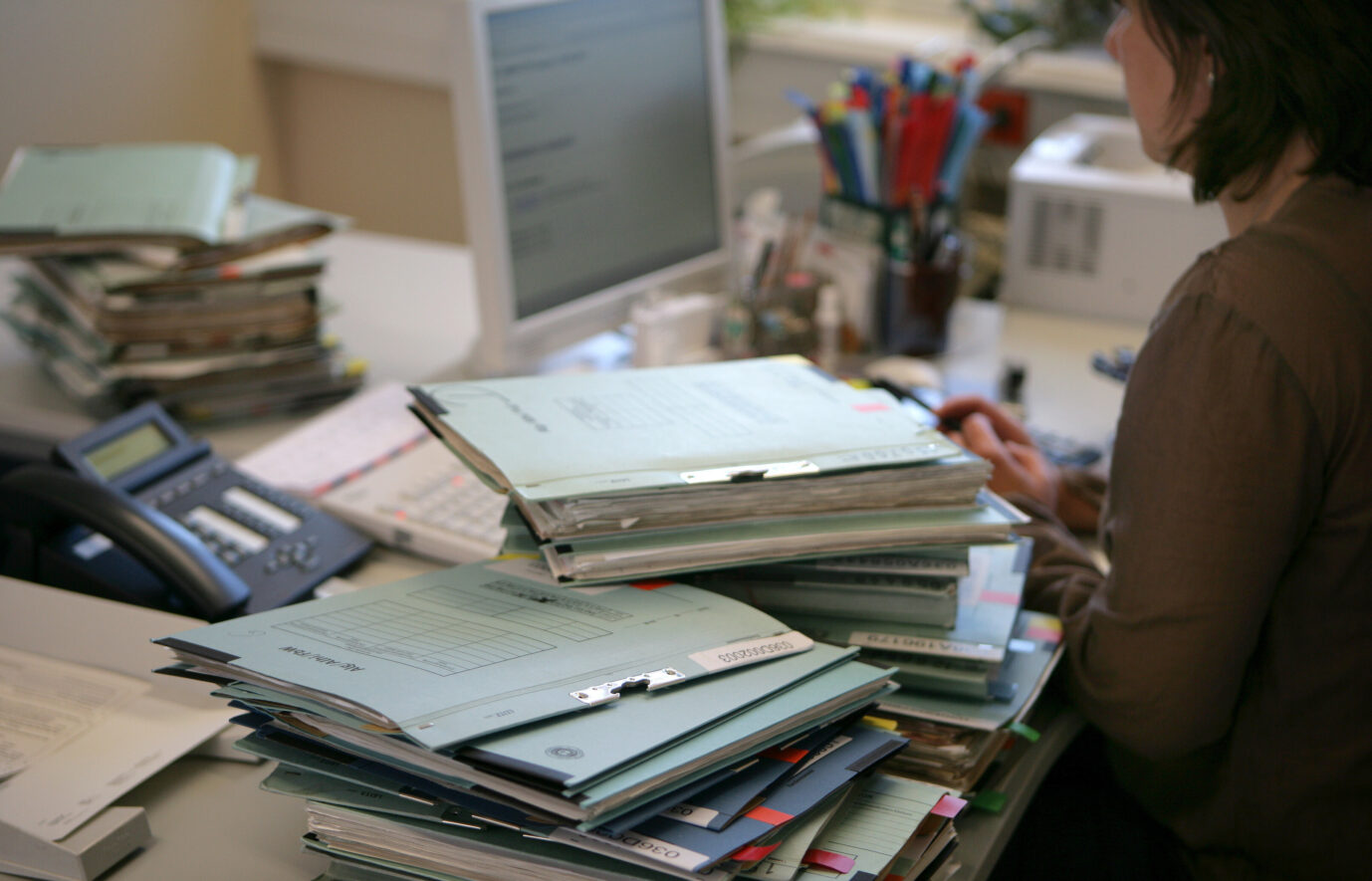Auf einem Büroschreibtisch liegt ein Stapel von Aktenordnern – Sinnbild einer überbordenden Bürokratie