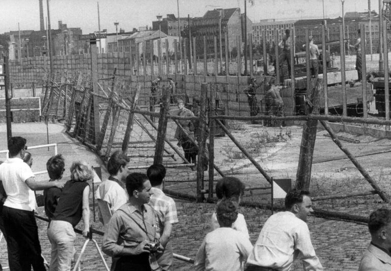 Auf einem alten Schwarz-Weiß-Foto sieht man Passanten, die Ostberliner Grenzsoldaten bei der Errichtung von neuen Grenzanlagen in Berlin zuschauen – Mauerbau in der DDR eben