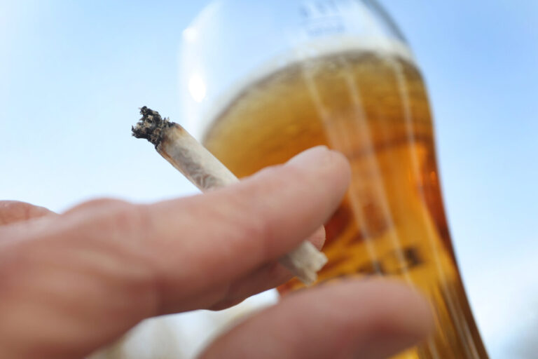 Eine Hand hält einen angezündeten Joint neben einem Bierglas. Im Hintergrund ist der blaue Himmel zu sehen. Das Bild steht im Zusammenhang mit der Diskussion um die Cannabis-Legalisierung und die geplanten Einschränkungen beim Alkoholkonsum in Deutschland. Hand mit Joint und Bierglas: Seit April 2023 ist Cannabis teilweise legal – nun soll Alkohol stärker eingeschränkt werden. Foto: picture alliance/dpa | Karl-Josef Hildenbrand