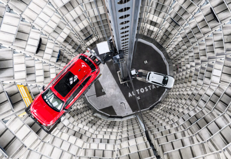 Zwei Autos werden in einem runden, hohen Fahrzeugturm bewegt, aufgenommen von oben. Das Bild zeigt ein automatisiertes Parksystem in der Autostadt Wolfsburg und steht für den Wandel der deutschen Autoindustrie. Fahrzeugturm in der Autostadt Wolfsburg: Der Standort gilt als Symbol der deutschen Autoindustrie – und ihres Ringens um die Zukunft. Foto: picture alliance/dpa | Julian Stratenschulte