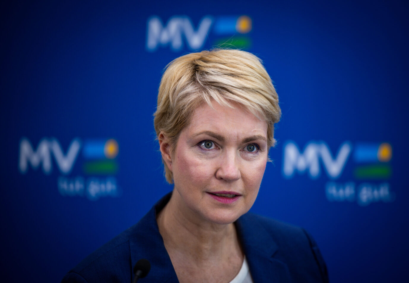 Eine Frau mit kurzem blondem Haar steht vor einer blauen Wand mit dem Logo des Landes Mecklenburg-Vorpommern. Sie blickt ernst in Richtung Kamera. Die Aufnahme zeigt die Ministerpräsidentin bei einer Pressekonferenz, bei der es um den Ausbau der Erstaufnahmeeinrichtung Stern Buchholz ging. Ministerpräsidentin von Mecklenburg-Vorpommern: Trotz sinkender Asylzahlen hält die Regierung am Ausbau der Erstaufnahme fest. Foto: picture alliance/dpa | Jens Büttner
