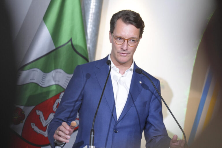 Ein mittelalter Herr in einem blauen Sakko mit weißem Hemd ohne Binder steht vor der grün-weiß-roten Flagge des Landes Nordrhein-Westfalen. Ministerpräsident Hendrik Wüst gestikuliert vor zwei Richtmikrofonen. Der Christdemokrat regiert das Land gemeinsam mit den Grünen. Nordrhein-Westfalens Ministerpräsident Hendrik Wüst: Die schwarz-grüne Landesregierung hält an der Förderung des Grimme-Instituts fest. Foto: picture alliance / Flashpic | Jens Krick