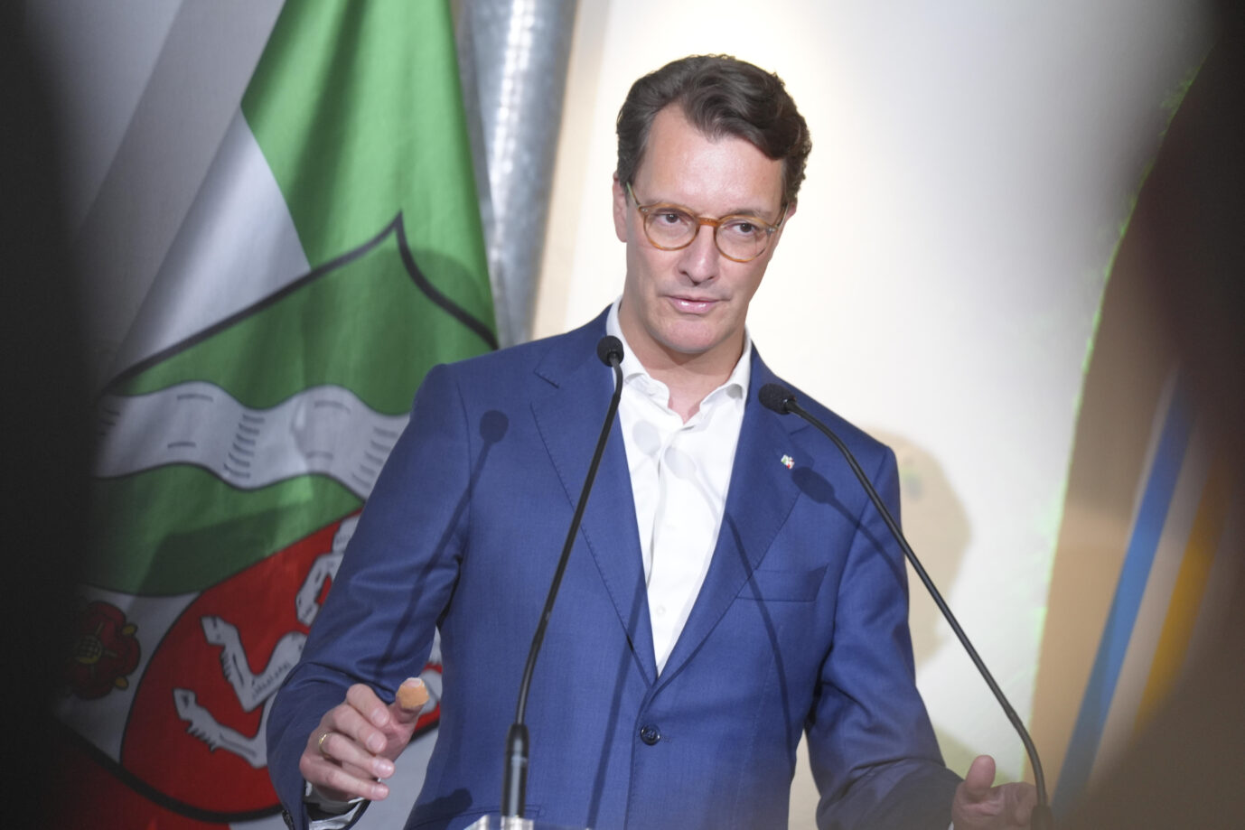 Ein mittelalter Herr in einem blauen Sakko mit weißem Hemd ohne Binder steht vor der grün-weiß-roten Flagge des Landes Nordrhein-Westfalen. Ministerpräsident Hendrik Wüst gestikuliert vor zwei Richtmikrofonen. Der Christdemokrat regiert das Land gemeinsam mit den Grünen. Nordrhein-Westfalens Ministerpräsident Hendrik Wüst: Die schwarz-grüne Landesregierung hält an der Förderung des Grimme-Instituts fest. Foto: picture alliance / Flashpic | Jens Krick