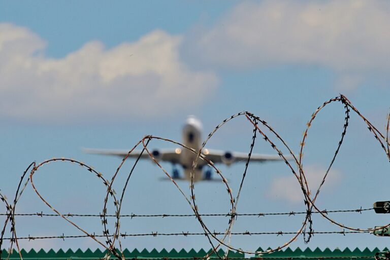 Stacheldrahtzaun mit startendem Flugzeug im Hintergrund, Symbol für Abschiebung und Asylrecht in Deutschland. Stacheldraht am Flughafenzaun: Trotz Vorstrafen blieb der Eritreer von Horb durch subsidiären Schutz in Deutschland. Foto: picture alliance / Daniel Kubirski | Daniel Kubirski