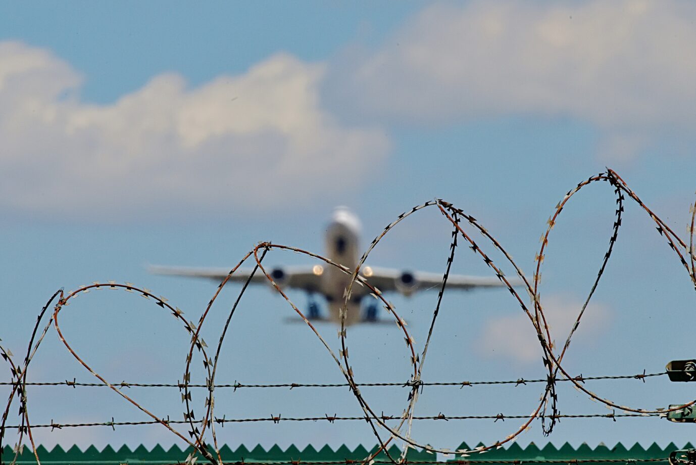 Stacheldrahtzaun mit startendem Flugzeug im Hintergrund, Symbol für Abschiebung und Asylrecht in Deutschland. Stacheldraht am Flughafenzaun: Trotz Vorstrafen blieb der Eritreer von Horb durch subsidiären Schutz in Deutschland. Foto: picture alliance / Daniel Kubirski | Daniel Kubirski