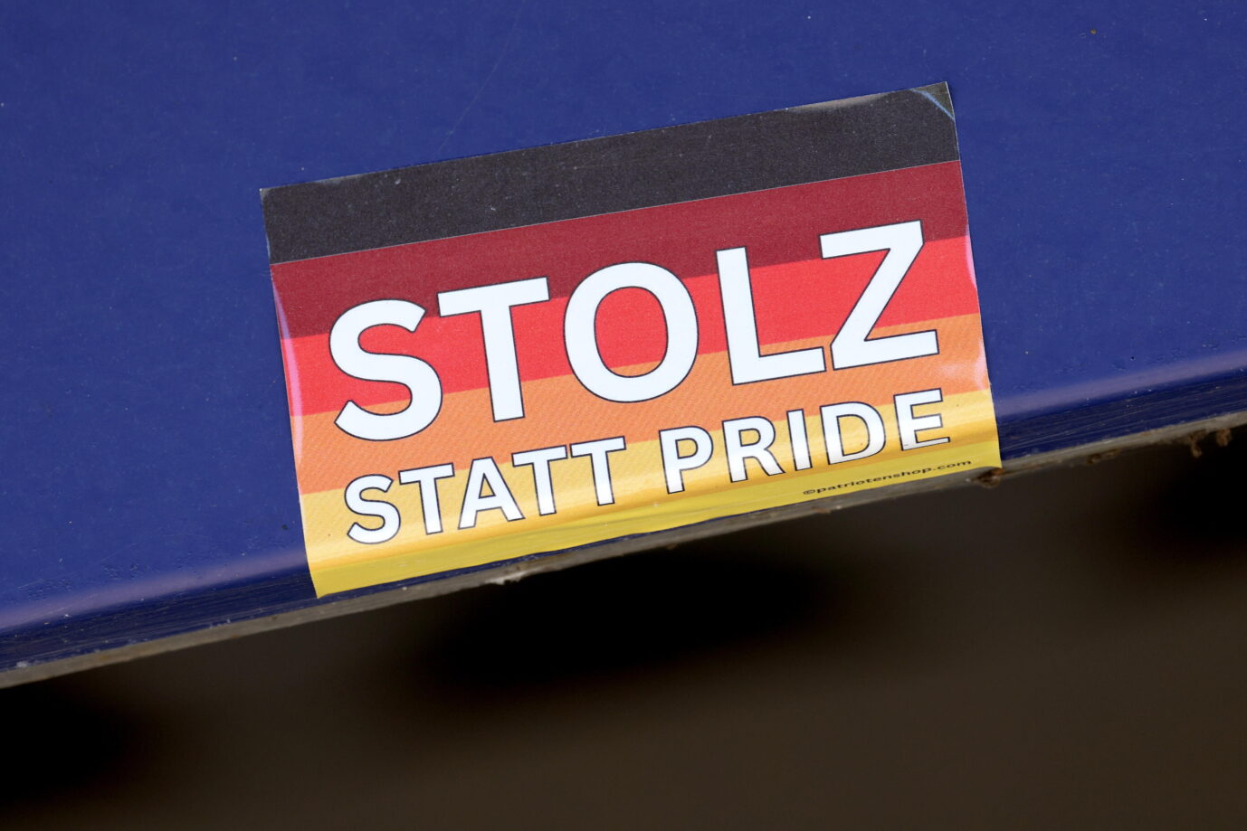 Ein rechteckiger Aufkleber mit der Deutschlandflagge im Hintergrund und dem Text „Stolz statt Pride“ klebt auf einer blauen Oberfläche. Der Sticker stammt aus dem Onlineversand „patriotenshop.com“ und wurde auch bei der Aktion gegen die Linkspartei in Hofgeismar eingesetzt. Sticker mit Deutschlandfarben und der Aufschrift „Stolz statt Pride“: Ein Motiv, das auch am Schaukasten der Linken in Hofgeismar verwendet wurde. Nun ermittelt der Staatsschutz. Foto: picture alliance / Rene Traut Fotografie | Rene Traut