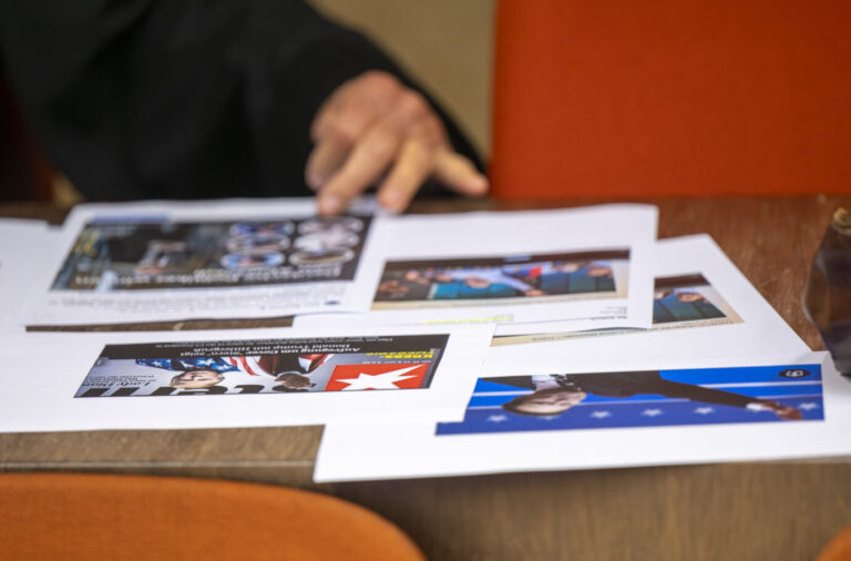 Ein Mann ordnet auf einem Tisch mehrere ausgedruckte Fotos und Dokumente, im Hintergrund orangefarbene Stühle. Zu sehen ist Verteidiger Peter Solloch, der im Verfahren gegen AfD-Europaabgeordneten Petr Bystron tätig ist. Thema: Anklage wegen Verwendens von Kennzeichen verfassungswidriger Organisationen. Anwalt Peter Solloch vor Prozessbeginn: Er verteidigt den AfD-Politiker Petr Bystron, der zum Termin nicht erschien. Foto: picture alliance/dpa | Peter Kneffel