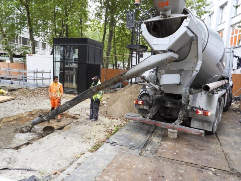 Auf einem öffentlichen Platz stehen Bauarbeiter und ein großer Zementmischer – daraus ragt ein Rohr und aus diesem fließt Beton auf eine hohle Stelle – dort sollte ursprünglich eine Toilette gebaut werden