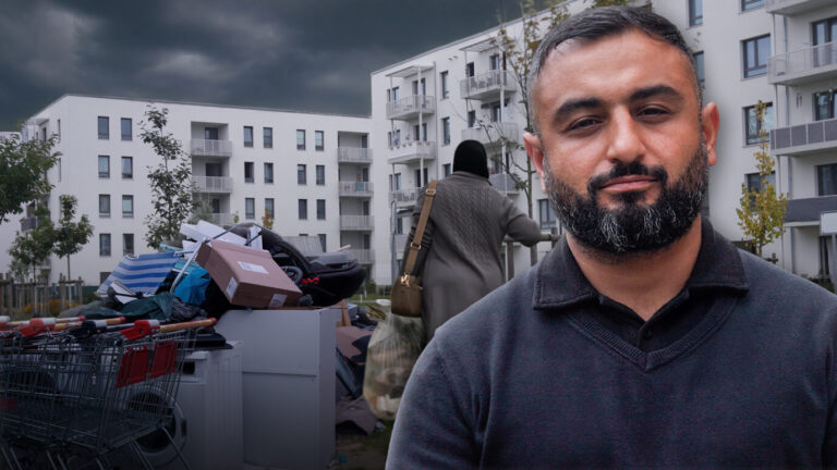 Ein Mann steht vor modernen weißen Wohnhäusern, im Vordergrund liegen Müll und Einkaufswagen. Im Hintergrund eine Frau mit Kopftuch. Die Aufnahme zeigt Korey Badem vor der Unterkunft am Hassoweg in Alt-Glienicke, wo sich Anwohner über zunehmende Spannungen beklagen. Familie Badem zieht von Kreuzberg in den Berliner Süden, auf der Suche nach Ruhe. Statt Idylle findet sie eine Unterkunft für Migranten vor der Haustür. Doch der Vater will sich nicht verdrängen lassen. Die JUNGE FREIHEIT auf Ortsbesichtigung. Korey Badem vor den Neubauten am Hassoweg: Aus dem Traum vom ruhigen Wohnen wurde ein Albtraum. Foto: JF