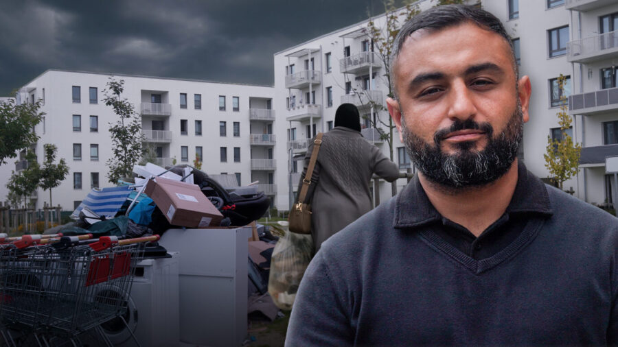 Ein Mann steht vor modernen weißen Wohnhäusern, im Vordergrund liegen Müll und Einkaufswagen. Im Hintergrund eine Frau mit Kopftuch. Die Aufnahme zeigt Korey Badem vor der Unterkunft am Hassoweg in Alt-Glienicke, wo sich Anwohner über zunehmende Spannungen beklagen. Familie Badem zieht von Kreuzberg in den Berliner Süden, auf der Suche nach Ruhe. Statt Idylle findet sie eine Unterkunft für Migranten vor der Haustür. Doch der Vater will sich nicht verdrängen lassen. Die JUNGE FREIHEIT auf Ortsbesichtigung. Korey Badem vor den Neubauten am Hassoweg: Aus dem Traum vom ruhigen Wohnen wurde ein Albtraum. Foto: JF