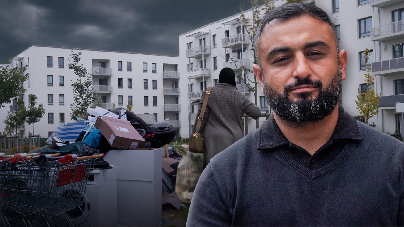 Ein Mann steht vor modernen weißen Wohnhäusern, im Vordergrund liegen Müll und Einkaufswagen. Im Hintergrund eine Frau mit Kopftuch. Die Aufnahme zeigt Korey Badem vor der Unterkunft am Hassoweg in Alt-Glienicke, wo sich Anwohner über zunehmende Spannungen beklagen. Familie Badem zieht von Kreuzberg in den Berliner Süden, auf der Suche nach Ruhe. Statt Idylle findet sie eine Unterkunft für Migranten vor der Haustür. Doch der Vater will sich nicht verdrängen lassen. Die JUNGE FREIHEIT auf Ortsbesichtigung. Korey Badem vor den Neubauten am Hassoweg: Aus dem Traum vom ruhigen Wohnen wurde ein Albtraum. Foto: JF