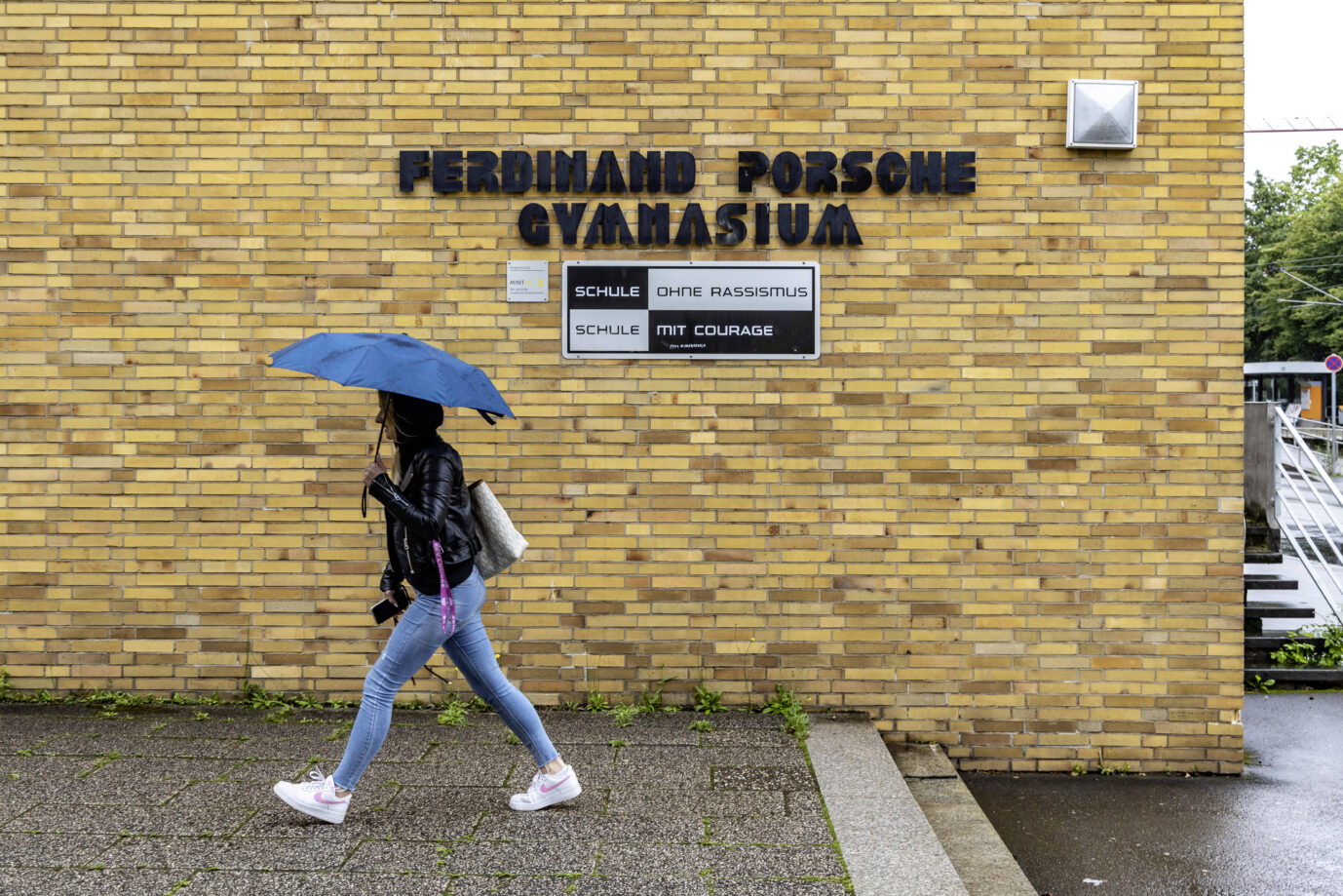 Eine junge Frau in schwarzer Jacke und mit blauem Regenschirm läuft am Schriftzug des Ferdinand-Porsche-Gymnasiums in Stuttgart-Zuffenhausen vorbei