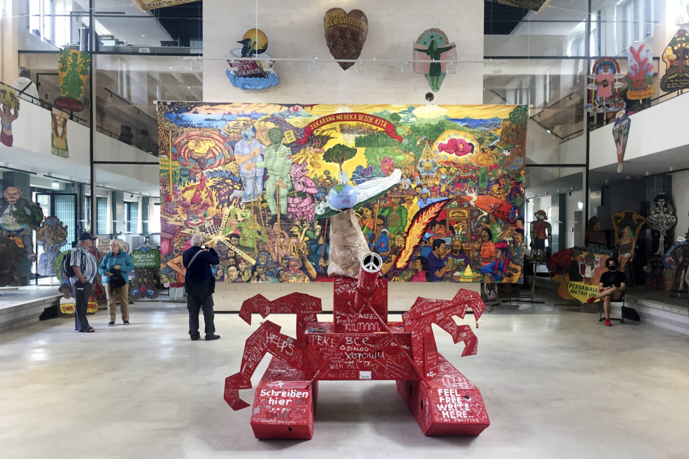 In einer Halle stehen verschiedene Kunstwerke, vorne eine rote Skulptur und im Hintergrund das „People’s Justice“-Banner eines indonesischen Künstlerkollektivs, welches als antisemitisch kritisiert wurde – auf der Documenta 15
