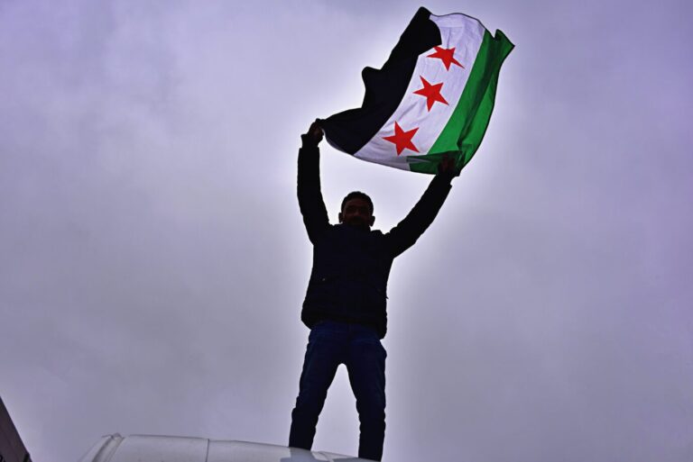 Ein Mann steht auf einem Fahrzeug und hält die syrische Oppositionsflagge mit drei roten Sternen über dem Kopf, aufgenommen bei einer Kundgebung in Frankfurt nach dem Sturz des Assad-Regimes im Dezember 2024. Die Syrer wollten zurückkehren. Ein Mann schwenkt in Frankfurt die syrische Oppositionsflagge: Nach Assads Sturz blieb die Rückkehrwelle aus. Foto: IMAGO / Daniel Kubirski