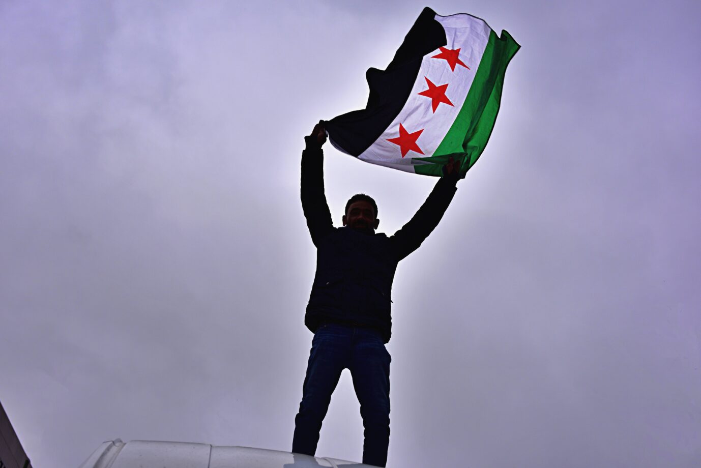 Ein Mann steht auf einem Fahrzeug und hält die syrische Oppositionsflagge mit drei roten Sternen über dem Kopf, aufgenommen bei einer Kundgebung in Frankfurt nach dem Sturz des Assad-Regimes im Dezember 2024. Die Syrer wollten zurückkehren. Ein Mann schwenkt in Frankfurt die syrische Oppositionsflagge: Nach Assads Sturz blieb die Rückkehrwelle aus. Foto: IMAGO / Daniel Kubirski