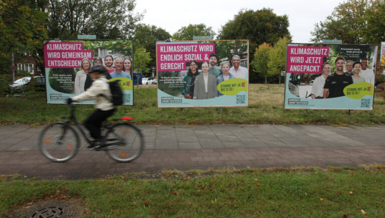 Ein Radfahrer fährt über einen Bürgersteig, im Hintergrund sind Wahlplakate zu sehen, die für den Hamburger „Zukunftsentscheid“ werben – demnach muß die Stadt beinahe komplett auf erneuerbare Energie setzen