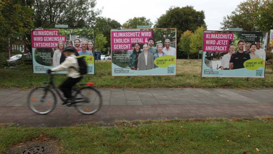 Ein Radfahrer fährt über einen Bürgersteig, im Hintergrund sind Wahlplakate zu sehen, die für den Hamburger „Zukunftsentscheid“ werben – demnach muß die Stadt beinahe komplett auf erneuerbare Energie setzen