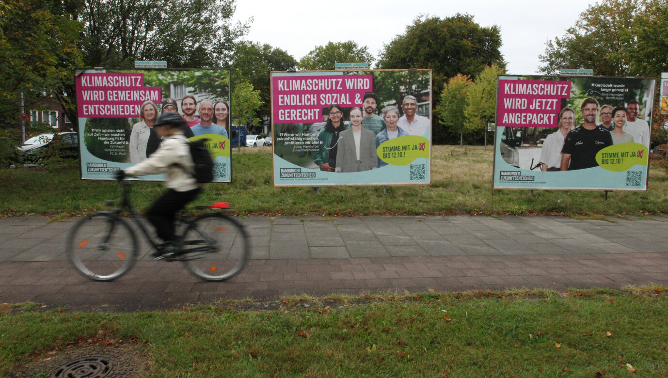 Ein Radfahrer fährt über einen Bürgersteig, im Hintergrund sind Wahlplakate zu sehen, die für den Hamburger „Zukunftsentscheid“ werben – demnach muß die Stadt beinahe komplett auf erneuerbare Energie setzen