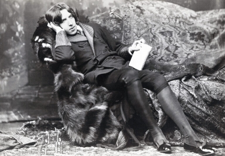 Ein altes schwarz-weißes Foto zeigt den Schriftsteller Oscar Wilde, wie er auf einem Sessel sitzt und ein Buch in der Hand hält