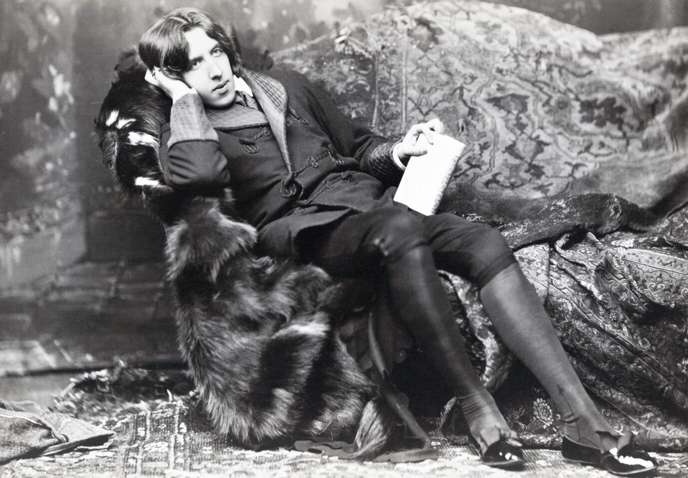 Ein altes schwarz-weißes Foto zeigt den Schriftsteller Oscar Wilde, wie er auf einem Sessel sitzt und ein Buch in der Hand hält
