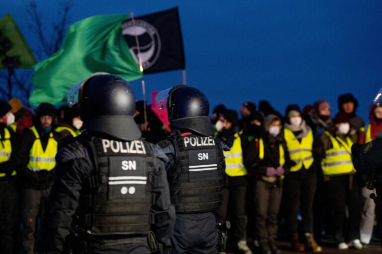 Polizisten in Schutzkleidung stehen in der Dunkelheit einer dicht gedrängten Gruppe von Demonstranten gegenüber, von denen viele Warnwesten tragen und Fahnen hochhalten, darunter eine grüne und eine schwarz-rote. Polizei und Gegendemonstranten vor dem AfD-Parteitag in der WT Energiesysteme Arena in Riesa am 11. Januar 2025, bei dem blockierte Zufahrten den Beginn verzögerten. Demonstrationen gegen den AfD-Parteitag in Riesa: Auch in Gießen wollen Linksextreme blockieren. Foto: picture alliance / PIC ONE | Bastian Stock