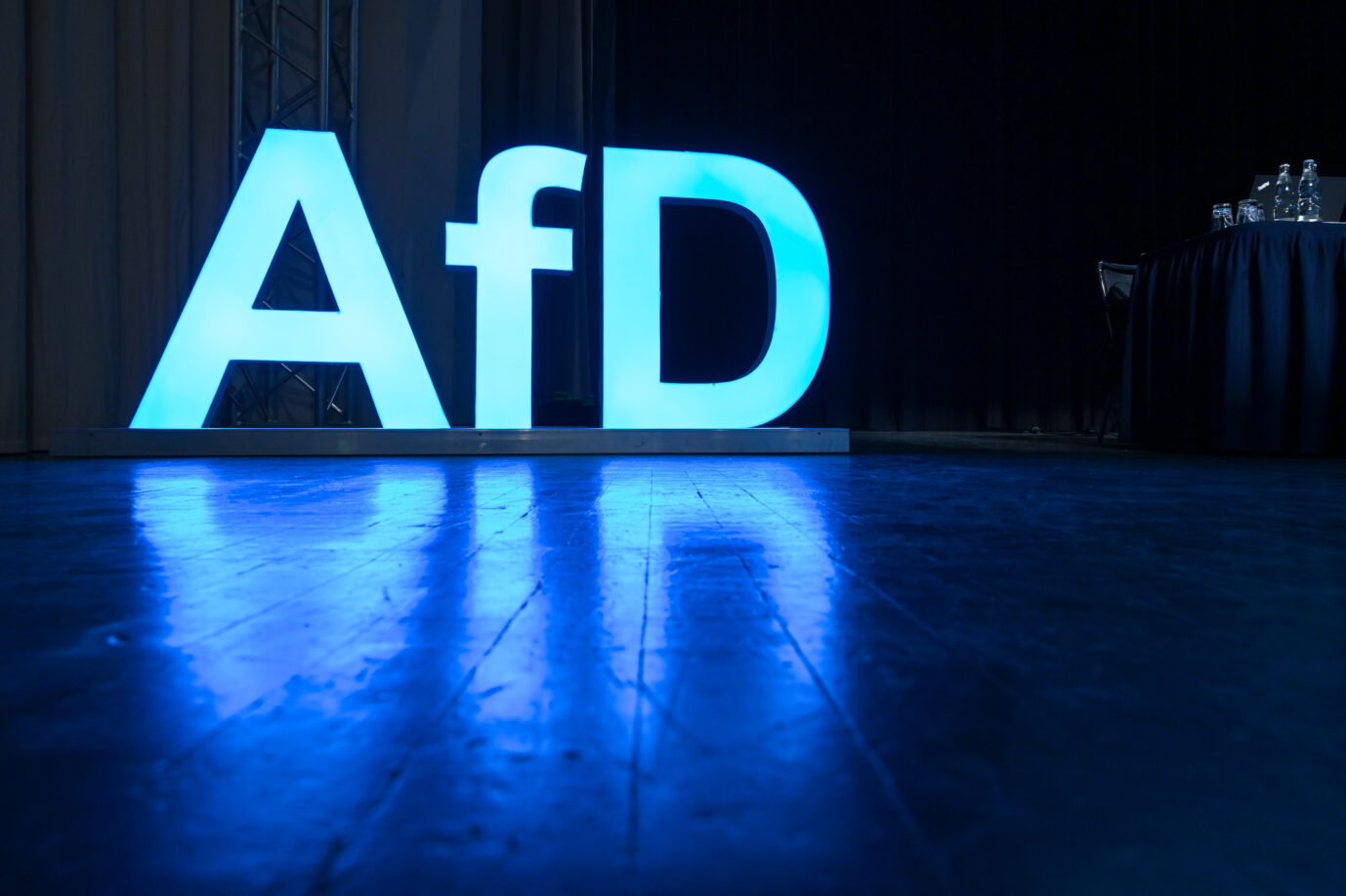 Großes, blau leuchtendes AfD-Logo auf einer Bühne. Die Buchstaben spiegeln sich auf dem dunklen Boden, rechts steht ein Tisch mit mehreren Wasserflaschen und Gläsern. Die Umgebung wirkt wie ein Veranstaltungssaal mit Verdunkelung im Hintergrund. AfD- Schriftzug und das AfD-Logo auf der Bühne im Versammlungssaal. Es werden die Kandidaten für die Landtagswahl 2026 aufgestellt. Leuchtlogo der AfD auf einem Parteitag: Für die AfD-Jugend gibt es divergierende Namenswünsche. Foto: picture alliance/dpa | Heiko Rebsch