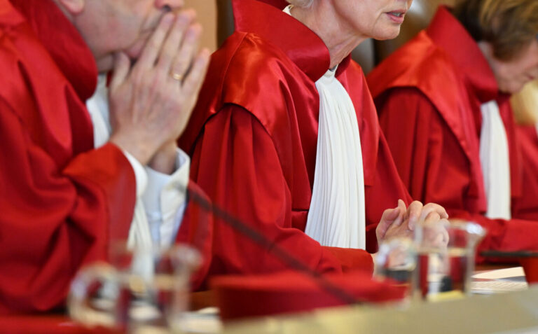 Richter in roten Roben sitzen nebeneinander im Sitzungssaal des Bundesverfassungsgerichts, ihre Hände gefaltet oder aneinandergelegt; die Aufnahme zeigt den oberen Körperbereich der Richter in Nahansicht. Richter des Bundesverfassungsgerichts in Karlsruhe: Entscheidung zu Polizeizugriff in Berliner Unterkunft. Foto: picture alliance/dpa | Uli Deck