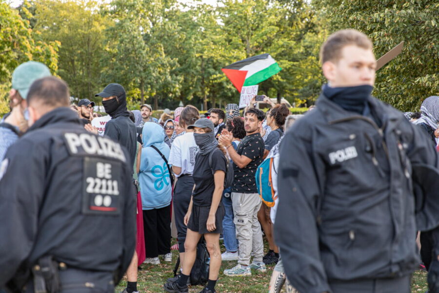 Vorne stehen Polizeibeamte in Uniform und schauen in die Ferne, hinten stehen Pro-Palästina-Demonstranten und schwenken Palästina Fahnen