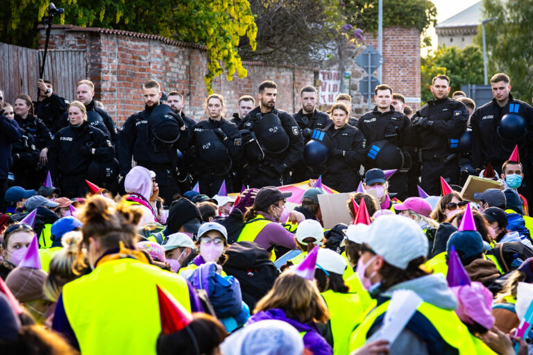 Polizisten stehen vor einer großen Gruppe sitzender Demonstranten in bunten Westen und mit Partyhütchen bei einer Aktion des Bündnisses Widersetzen in Demmin. Das Bündnis organisiert regelmäßig Proteste gegen Veranstaltungen der AfD. Polizeikette vor Sitzblockierern bei einer Aktion des Bündnisses Widersetzen in Demmin. Foto: Santiago Rodríguez / Widersetzen / CC BY-SA 4.0