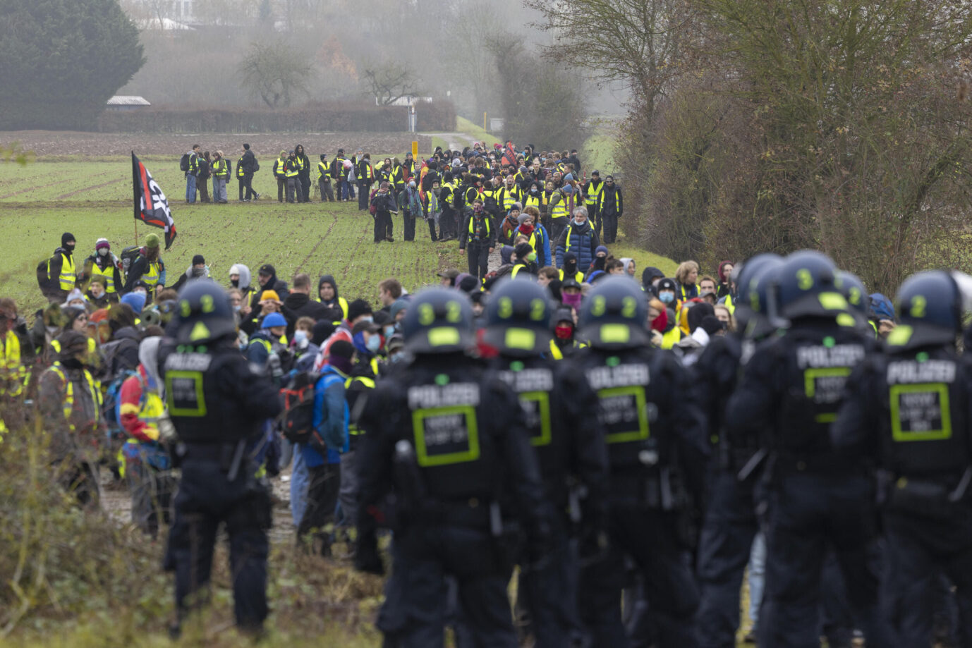 Polizisten in Schutzuniformen stehen herum, im Hintergrund sieht man linksextreme Demonstranten, Gießen