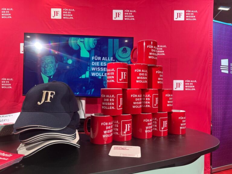 Rote Tassen stapeln sich neben blauen Mützen, dahinter groß das Logo der JUNGEN FREIHEIT - der JF-Stand bei der Buchmesse Seitenwechsel