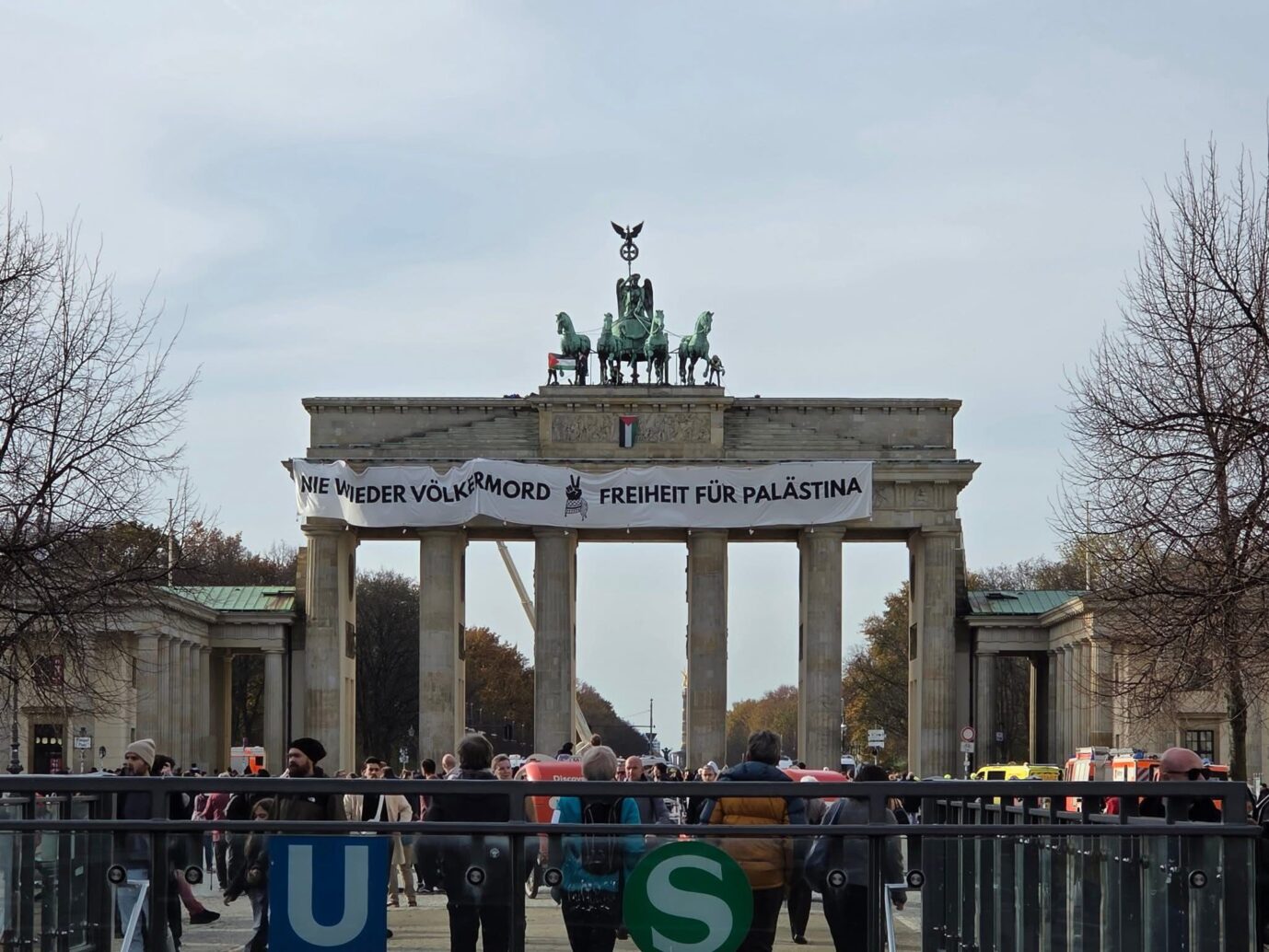 Blick auf das Brandenburger Tor mit einem großen, weißen Banner über dem Mittelbau, darauf die Aufschrift „Nie wieder Völkermord – Freiheit für Palästina“. Drei Personen standen zuvor oben auf dem Tor, wurden aber bereits von der Polizei in Gewahrsam genommen; das Banner ist inzwischen entfernt. Im Vordergrund dichter Fußverkehr nahe dem Eingang zur U- und S-Bahn. Drei Personen auf dem Brandenburger Tor mit einem Spannbanner: „Nie wieder Völkermord – Freiheit für Palästina“. Foto: privat