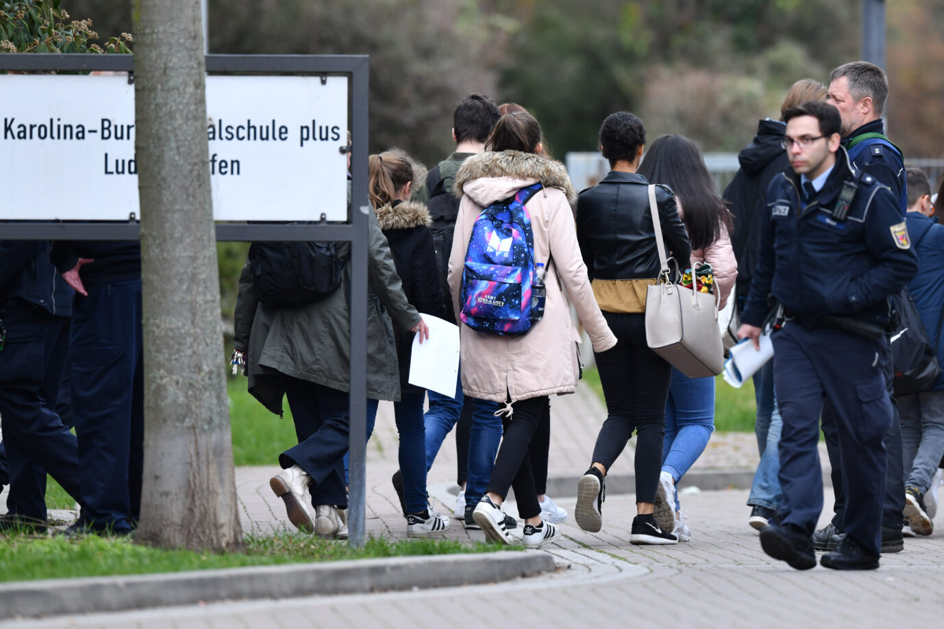 Schüler gehen an einem Schild der Karolina-Burger-Realschule in Ludwigshafen vorbei, daneben stehen Polizisten. Jetzt haben sich die Lehrer der Schule in einem Brandbrief hilfesuchend an die Behörden gewandt