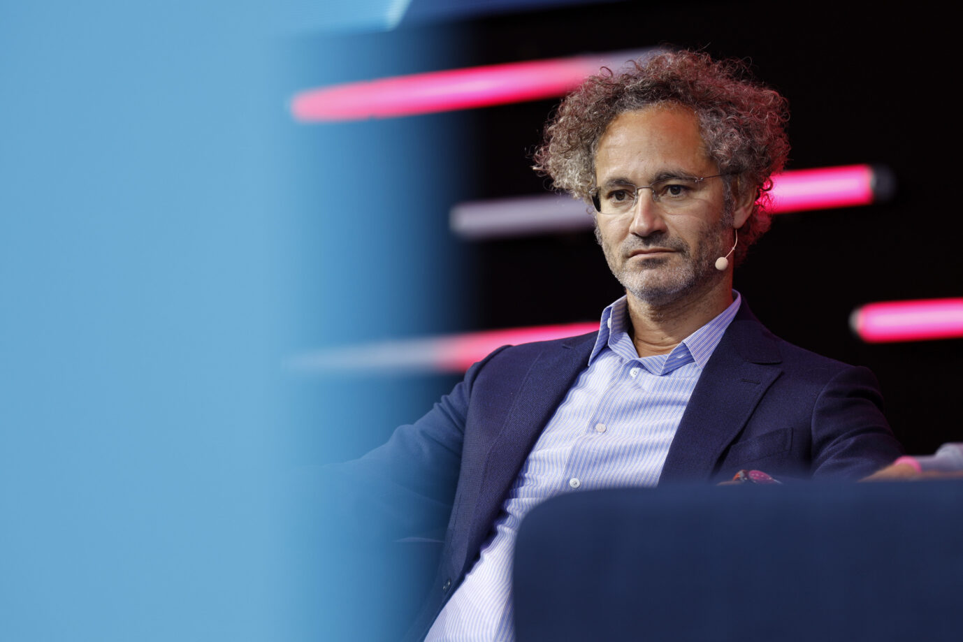 Alex Karp sitzt auf einer Bühne in einem Sessel, trägt Sakko und Hemd und blickt ernst nach vorn, im Hintergrund farbige Lichtstreifen. Palantir-CEO Alex Karp äußert sich im Handelsblatt-Interview kritisch zu Deutschlands technologischem Niedergang und zur Migrationspolitik. Palantir-CEO Alex Karp in Köln: hält nichts von Deutschlands Migrationspolitik. Foto: picture alliance / Geisler-Fotopress | Christoph Hardt/Geisler-Fotopres