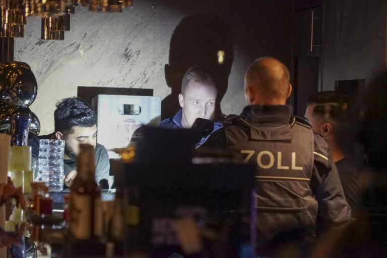 Zwei Zollbeamte kontrollieren im Schein einer Taschenlampe mehrere Männer hinter der Theke einer Shisha-Bar. Im dunklen Innenraum stehen Getränkeflaschen und Barzubehör, einer der Männer blickt auf Unterlagen, während der Lichtkegel die Szene scharf ausleuchtet. Gemeinsamer Einsatz von Zoll, Polizei, Finanzbehörde und Bezirksamt in mehreren Hamburger Bars am 7. Oktober 2022; dabei stellten die Kräfte Tabak sicher, löschten einen glühenden Kohleeimer und schlossen Lokale wegen erhöhter Kohlenmonoxidwerte. Zollbeamte überprüfen Mitarbeiter einer Shisha-Bar: Dutzende Rechtsverstöße. Foto: picture alliance /ABBfoto | -