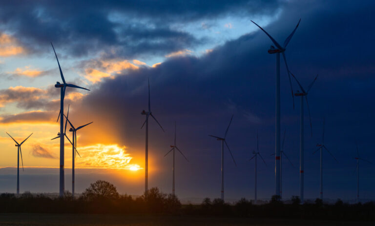 Vor einer untergehenden Sonne drehen sich Windkraftanlagen – Symbolbild für die Energiewende und ihre Windflauten und Dunkelflauten