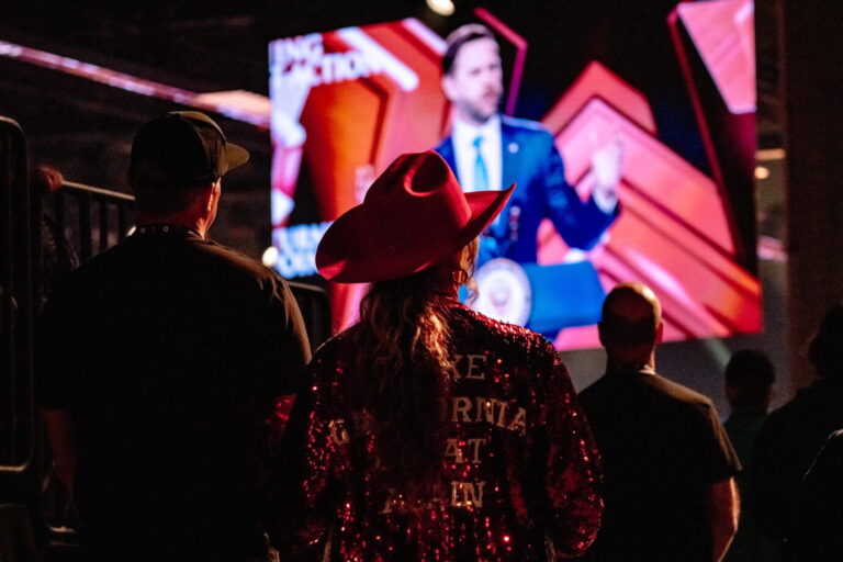 Teilnehmer stehen in einer Halle und blicken auf eine große Videoleinwand mit einem Redner auf der Bühne. Aufnahme vom AmericaFest der Studentenorganisation Turning Point USA in Phoenix, bei dem es zu offenen Konflikten innerhalb der MAGA-Bewegung kam. Besucher verfolgen beim AmericaFest von Turning Point USA eine Rede auf der Hauptbühne: Der innerkonservative Streit überschattet die Veranstaltung. Foto: picture alliance / Sipa USA | Laura Brett