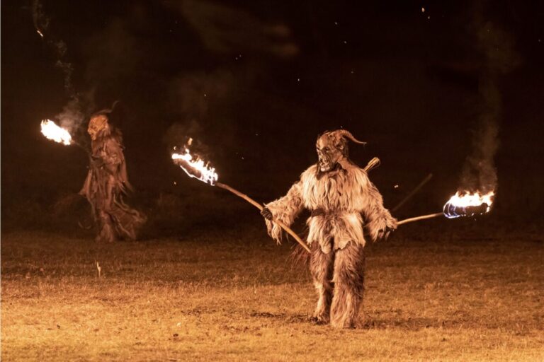 Zwei Perchten in zotteligen Kostümen stehen, brennende Fackeln in der Hand, in der Dunkelheit