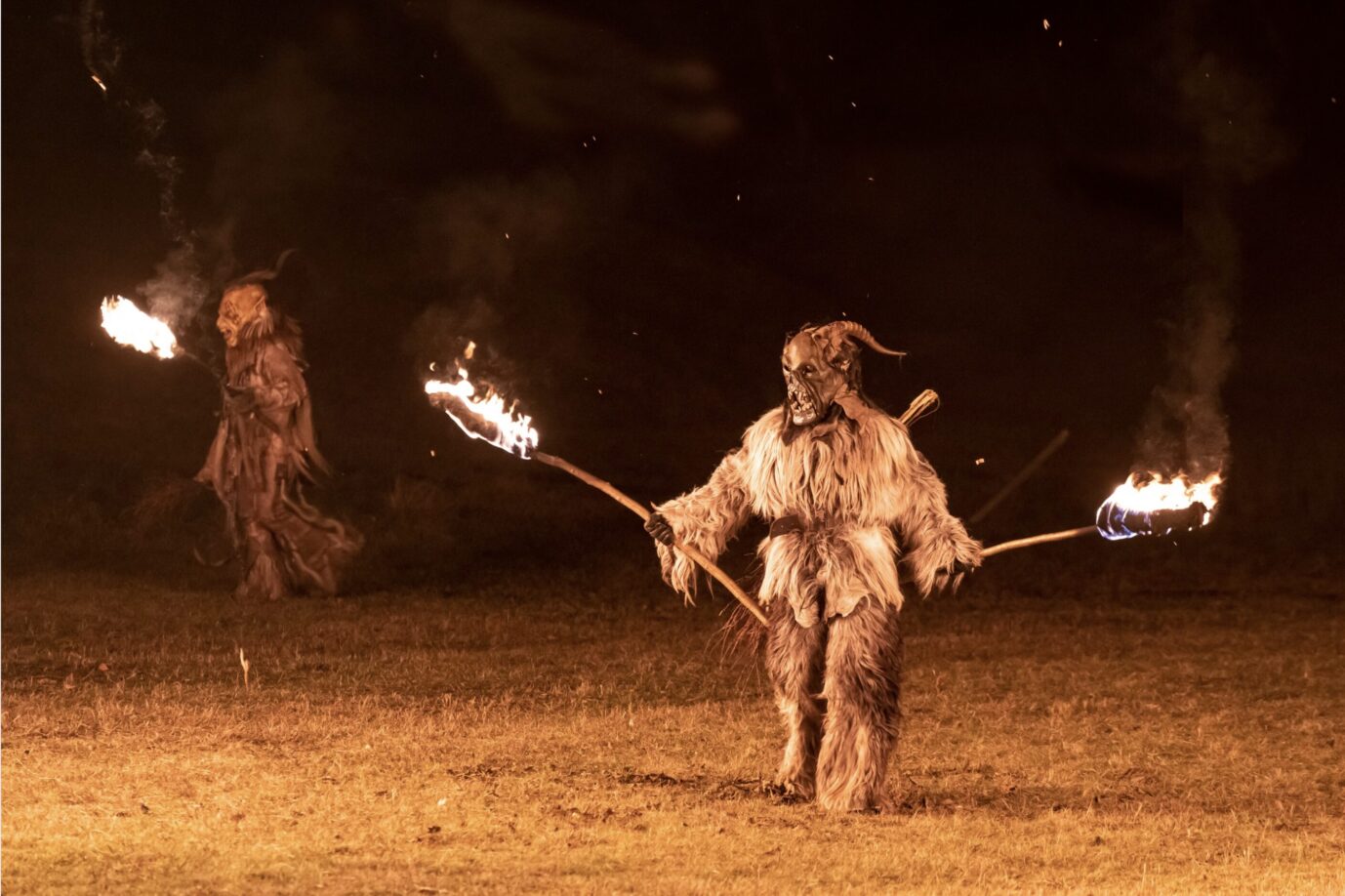 Zwei Perchten in zotteligen Kostümen stehen, brennende Fackeln in der Hand, in der Dunkelheit