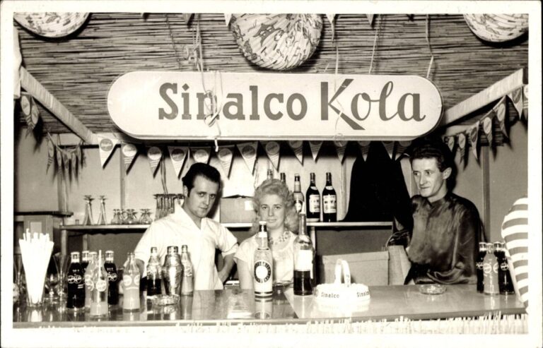 Auf einem alten Schwarz-Weiß-Bild sind Personen hinter einer Bar zu sehen, über ihnen hängt ein Schild von Sinalco-Kola