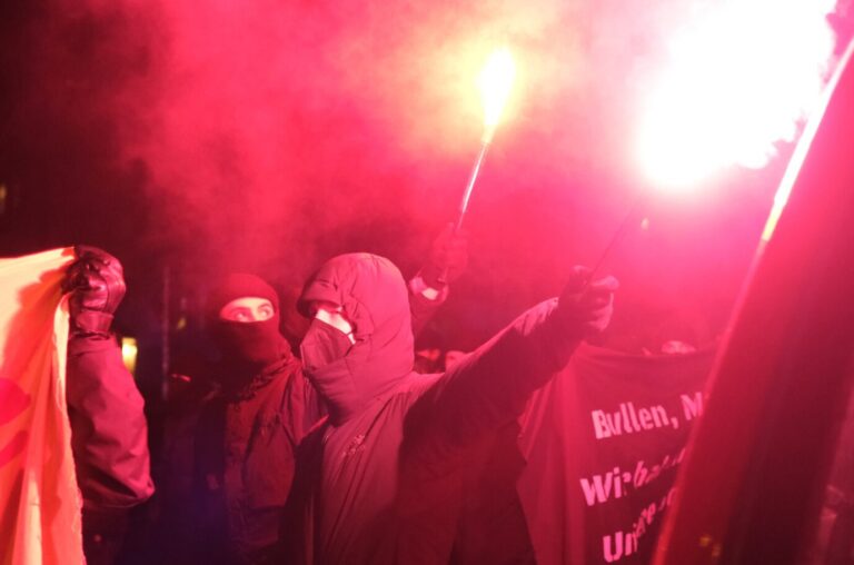 Mehrere vermummte Demonstranten gehen mit Transparenten und brennender Pyrotechnik durch eine Straße bei Dunkelheit. Die Aufnahme entstand bei einer linken Demonstration im Leipziger Stadtteil Connewitz am 12. Januar 2023 nach Wohnungsdurchsuchungen durch die Polizei. Teilnehmer einer linken Demonstration mit Pyrotechnik: Die „Roten Hilfe“ unterstützt nach eigenen Angaben auch Personen, die sich nach Polizeimaßnahmen als politisch verfolgt darstellen. Foto: picture alliance/dpa | Sebastian Willnow