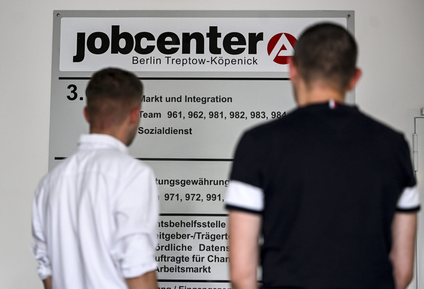Zwei Männer stehen mit dem Rücken zur Kamera vor einem Hinweisschild in einem Berliner Jobcenter. Das Schild weist auf das Jobcenter Berlin Treptow-Köpenick sowie auf Abteilungen wie Markt und Integration und den Sozialdienst hin und steht exemplarisch für den behördlichen Umgang mit Arbeitslosen und Leistungsbeziehern. Jobcenter in Berlin: Ukrainer erhielten Bürgergeld in zweistelliger Milliardenhöhe. Foto: picture alliance/dpa | Jens Kalaene