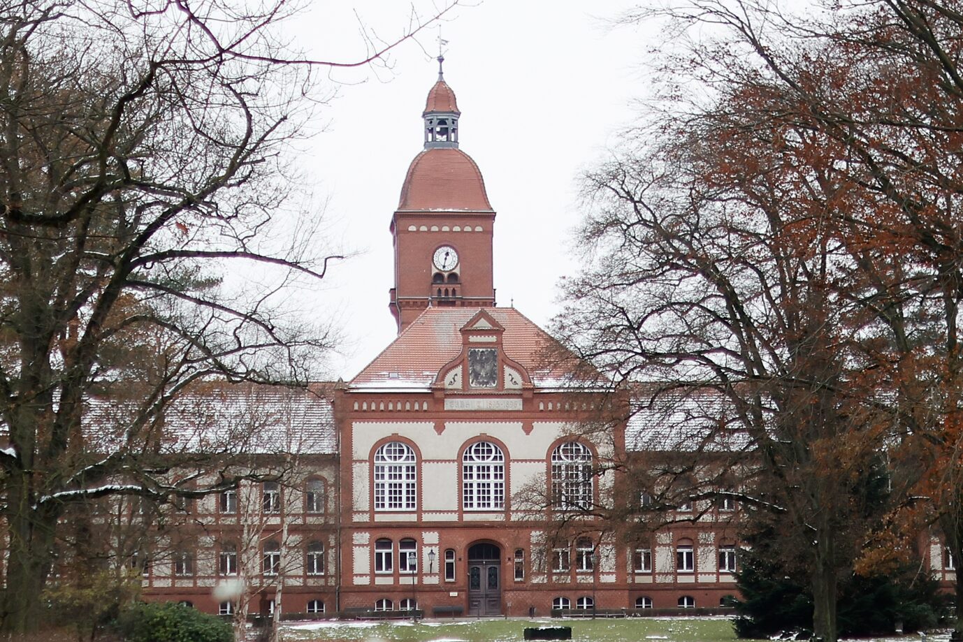 Backsteinbau mit Uhrturm und Haupteingang des Universitätsklinikums Ruppin-Brandenburg, umgeben von Bäumen im Winter. Großes Krankenhaus in Brandenburg, das Anfang 2024 die Schließung zweier Fachabteilungen ankündigte. Haupteingang des Universitätsklinikums Ruppin-Brandenburg in Neuruppin: Das Haus kündigte Anfang 2024 die Schließung zweier Fachabteilungen an. Foto: picture alliance/dpa | Carsten Koall
