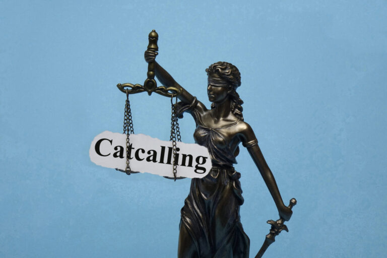 Bronzene Justitia-Statue mit Augenbinde hält eine Waage, an der ein Schild mit der Aufschrift „Catcalling“ hängt, vor blauem Hintergrund. Symbolbild zur Debatte über die strafrechtliche Einordnung von Anmachsprüchen und verbaler Belästigung. Justitia mit Waage und dem Schriftzug „Catcalling“: Hessen richtet eine neue Stelle bei der Generalstaatsanwaltschaft ein. Foto: picture alliance / ZB | Sascha Steinach
