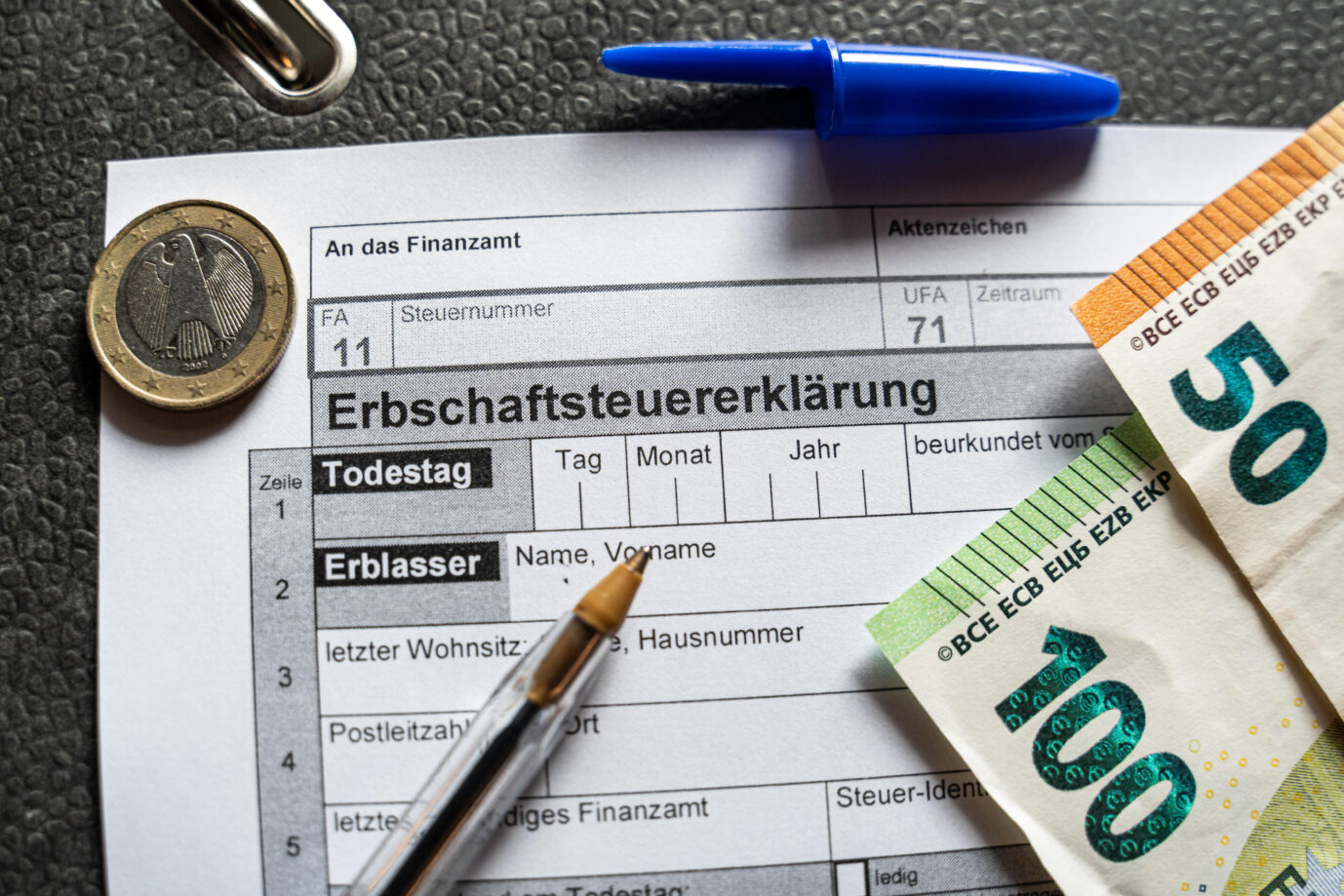 Formular zur Erbschaftssteuererklärung mit Euro-Banknoten, Münze und Stift auf einem Ordner. Symbolfoto für Nachlass, Finanzen und steuerliche Pflichten beim Erbe in Deutschland.