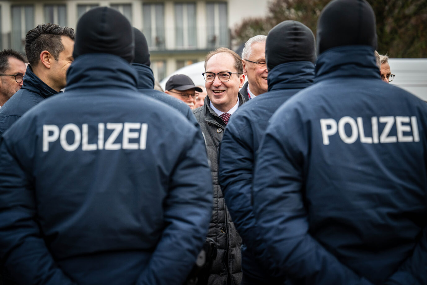 Bundesinnenminister Alexander Dobrindt steht im Freien zwischen mehreren Polizeibeamten in Einsatzkleidung. Die Beamten tragen Jacken mit der Aufschrift Polizei, im Hintergrund sind weitere Personen und Gebäude zu sehen. Bundesinnenminister Alexander Dobrindt (CSU) im Gespräch mit Polizeibeamten: Nach dem Anschlag auf das Berliner Stromnetz befürwortet er eine hohe Belohnung für Hinweise auf die Täter der Vulkangruppe.