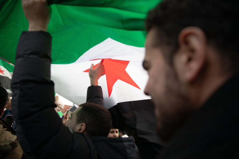 Mehrere Personen stehen dicht gedrängt unter einer großen Fahne der syrischen Opposition mit grün-weiß-schwarzen Streifen und rotem Stern. Die Aufnahme entstand bei einer Kundgebung syrischer Migranten am 7. Dezember 2025 auf dem Alexanderplatz in Berlin anläßlich des Jahrestags des Machtwechsels in Syrien. Syrische Demonstranten in Berlin: Trotz Debatte über Rückkehr entzieht das Bamf seltener Schutzstatus. Foto: picture alliance / ZUMAPRESS.com | Michael Kuenne