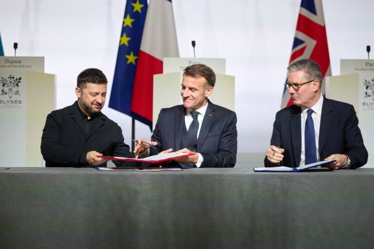 Drei Männer im Anzug sitzen an einem Tisch und unterzeichnen Dokumente, hinter ihnen stehen die Flaggen der Ukraine, Frankreichs und Großbritanniens. Aufnahme vom Treffen der Koalition der Willigen in Paris am 6. Januar 2026 mit Selenskyj, Macron und Merz zur Abstimmung weiterer Schritte im Ukraine-Krieg. Unterzeichnung in Paris: Der ukrainische Präsident Selenskyj, Frankreichs Staatspräsident Macron und Bundeskanzler Merz am Rande des Treffens der sogenannten Koalition der Willigen im Élysée-Palast. Foto: picture alliance / ZUMAPRESS.com | PRESIDENT OF UKRAINE