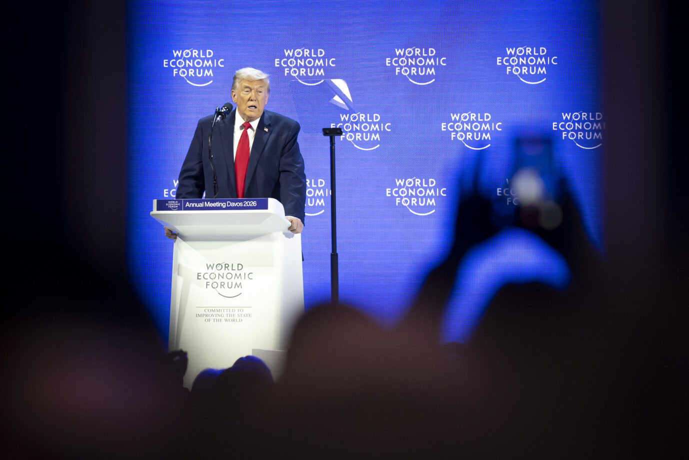 US-Präsident Donald Trump steht hinter einem Rednerpult mit der Aufschrift „World Economic Forum – Annual Meeting Davos 2026“ und spricht vor blauem Hintergrund mit WEF-Logo. Im Vordergrund sind unscharf die Silhouetten von Zuhörern und erhobenen Mobiltelefonen zu sehen, die die Rede fotografieren. Donald Trump spricht beim Weltwirtschaftsforum in Davos: Viele Freunde, wenige Feinde. Foto: picture alliance/KEYSTONE | LAURENT GILLIERON